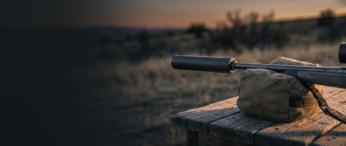 Suppressed rifle resting on a shooting bag on a wooden bench at dusk