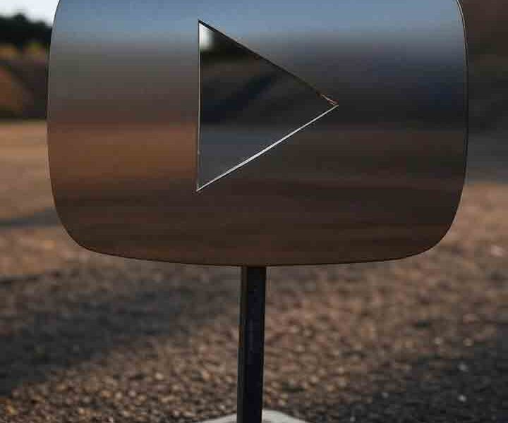 Steel target in the shape of a YouTube play button at an outdoor shooting range, with a metallic red triangle icon on a black square background.