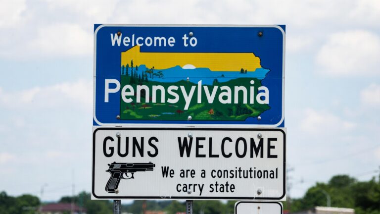 Blue Pennsylvania welcome sign with a scenic state illustration; below it a white sign that reads 'GUNS WELCOME' with a handgun graphic and 'We are a constitutional carry state'