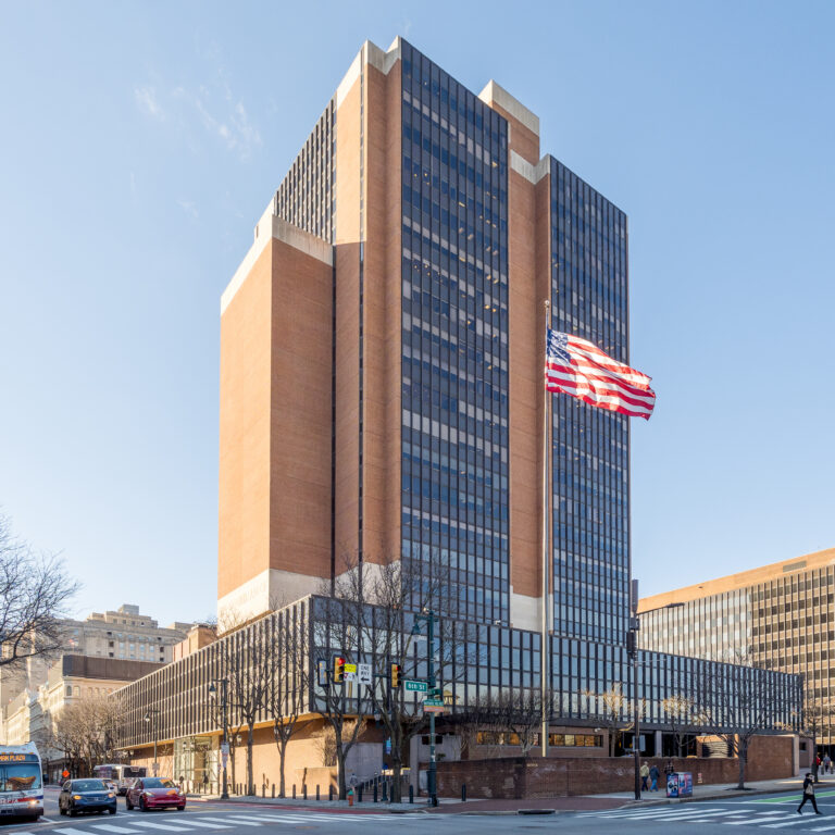 Tall brick-and-glass office building on a city street with a waving American flag at the corner.