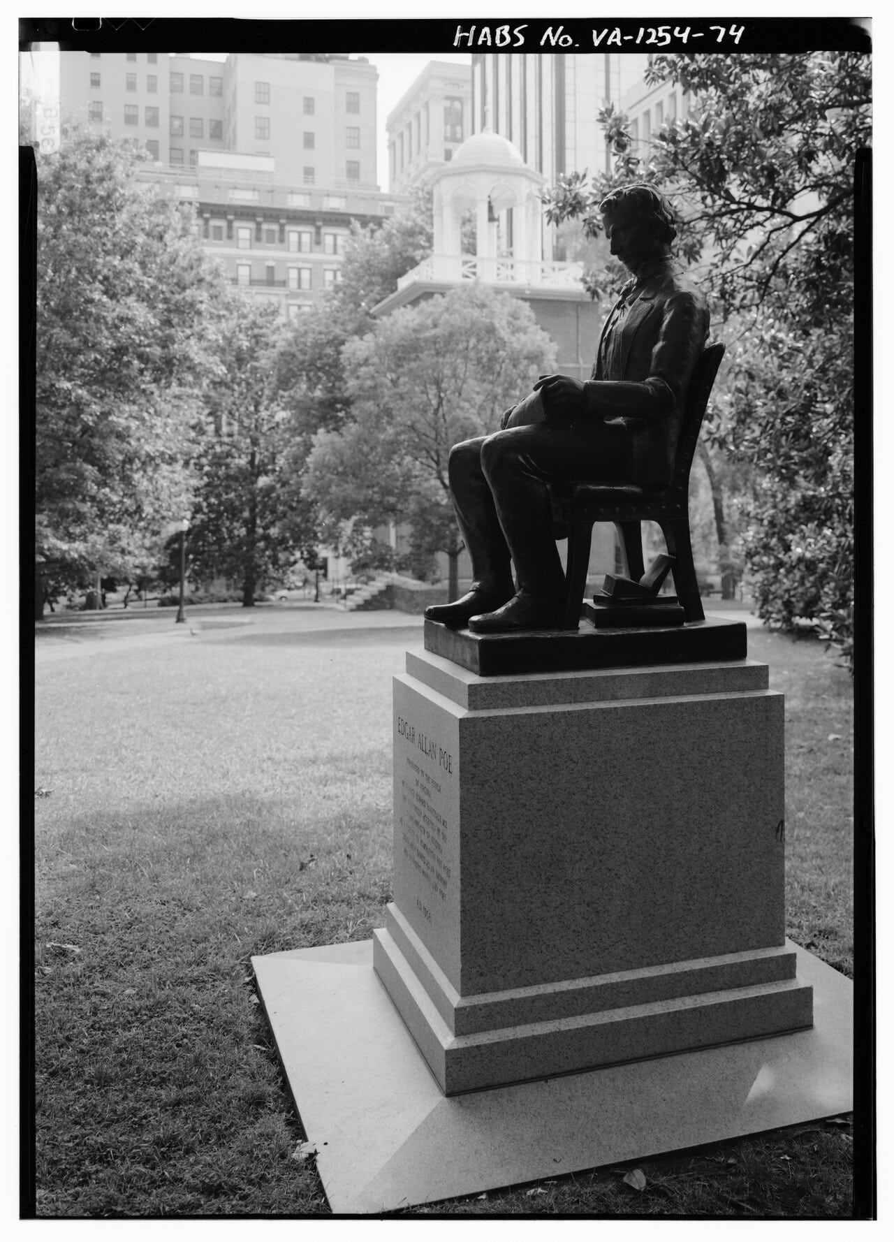 Bronze statue of Edgar Allan Poe seated in a chair on a stone pedestal in a city park, with trees and tall buildings in the background and a plaque on the pedestal.