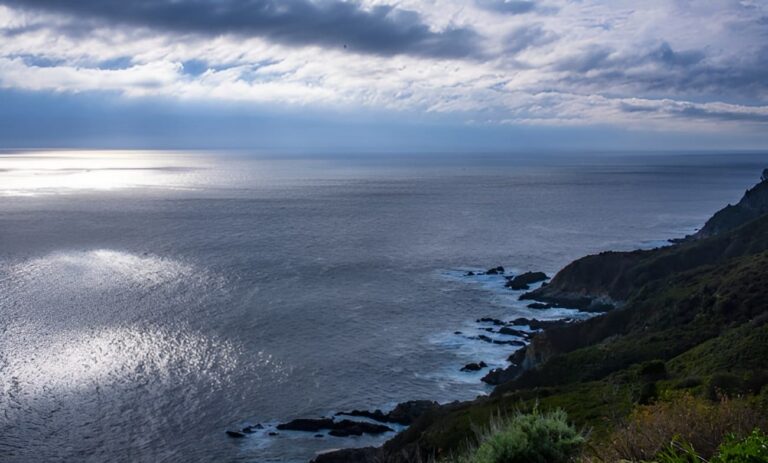Sunlight glinting on the calm sea along a rocky shoreline under cloudy skies. (Coastal cliff foreground)