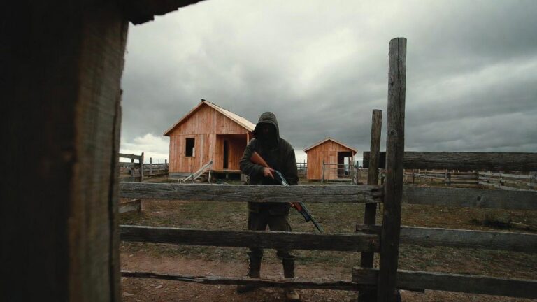 Hooded person behind a wooden fence holding a shotgun in a rural farmyard with wooden sheds and a cloudy sky.