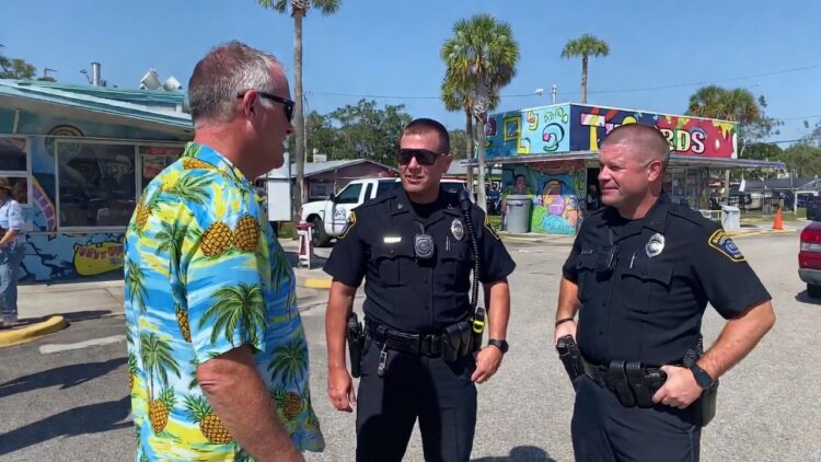 Police officers interviewing man on the side of the road - Freedom’s Lodge Three men stand and chat in a sunny parking lot; two police officers in black uniforms listen to a man in a bright pineapple-pattern shirt, with a colorful mural behind them.