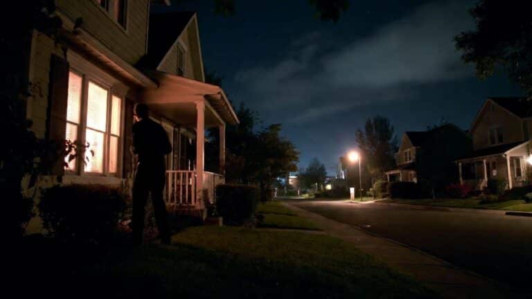 A man stands on a front yard at night, looking toward a warmly lit window of a house on a quiet suburban street.