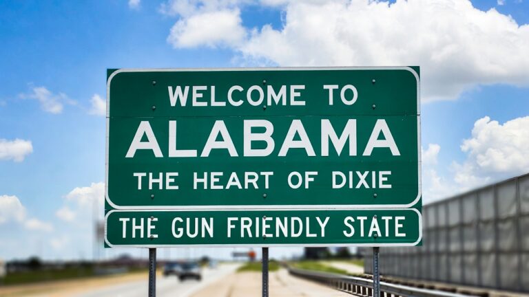 Green highway welcome sign reading 'Welcome to Alabama' with subtitles 'The Heart of Dixie' and 'The Gun Friendly State' on a clear day.